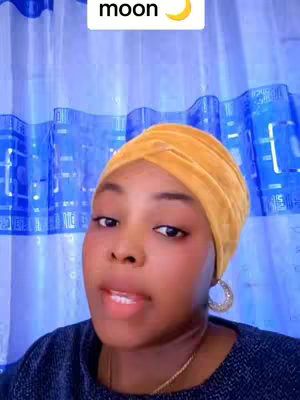 A woman with an orange head wrap and a navy blue top is shown in a selfie-style shot. She is looking slightly to the side and speaking, with a background of a blue curtain.