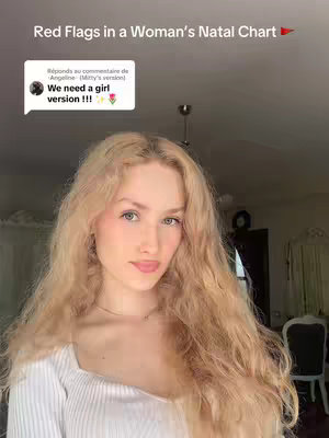 A young woman with long blonde hair and a white top is centered in the frame, facing the camera. Above her head, text overlay says 'Red Flags in a Woman's Natal Chart' next to a small red flag icon. A white speech bubble with a comment from another user is also present. The background is slightly blurred, with hints of a room and overhead lighting visible.