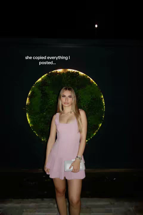 A young woman in a pink dress stands centered in front of a dark green backdrop with a circular design behind her. The woman has long blonde hair, holds a silver clutch, and wears a watch. Text overlayed at the top says, 'she copied everything I posted...'
