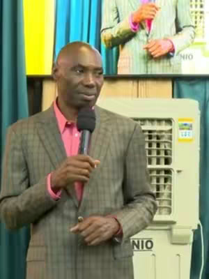 An individual, presumably a speaker or preacher, dressed in a suit coat and holding a microphone, stands in front of a pale green air conditioning unit. Behind him, another person in a suit can be seen in the background. The overall composition is formal, suggesting a public speaking or instructional context.