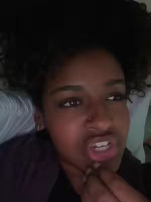 A close-up shot of a person with curly hair lying down and looking up at an angle. She has brown eyes and tan skin, with a nose ring, and her mouth is open as if she's speaking. The background is dark, suggesting a dim setting.