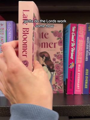 A hand holding a pink book in front of other books on a shelf. The hand is holding the book with the spine visible. The book on the right is titled "Late Bloomer" and the title of the book on the left is not visible. The words "gotta do the Lords work some how" are overlaid on the image.