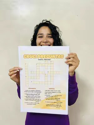 A woman with dark curly hair is smiling and holding up a white paper with a crossword puzzle on it. Behind her is a plain, light-colored wall.