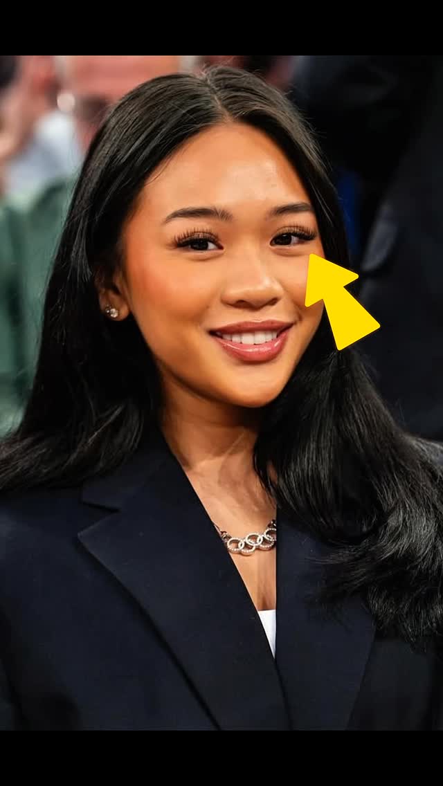 A portrait shot features Sunisa Lee, a young Asian woman with dark hair and a warm smile. A yellow arrow points to her eye, highlighting it. She is wearing a dark blazer over a white top and a silver necklace. The background is blurred, with indistinct people. The lighting is natural, and the overall composition is a close-up with a focus on her face.