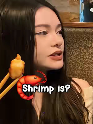 A woman with long black hair is centered, facing slightly to the side while seated at a table. In the leftmost foreground, a pair of chopsticks hold a piece of fried food with a golden crust. Below the food, a large red shrimp emoji and the text "Shrimp is?" are overlaid, in a white, bold font.