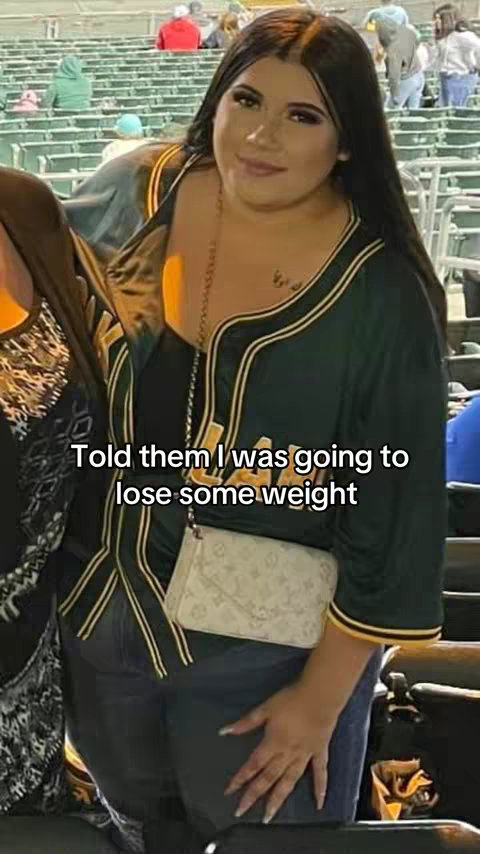 A woman in a dark green baseball jersey stands outdoors in front of stadium seating with a white purse and tattoos. Text overlayed on the image says, "Told them I was going to lose some weight".