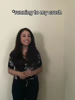 A young woman with long black hair stands with a slight smile in front of a beige wall. She wears a black top with floral patterns and black pants.