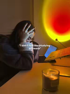 An interior shot showing a person resting their head on their hands on a desk. There is text overlayed on the image. Behind the person is a sunset lamp illuminating the room. There is a laptop and a glass on the table.
