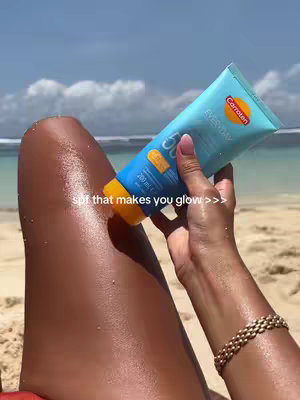 A person is holding a tube of sunscreen above a leg on a beach, presumably applying it. The leg appears sun-kissed. The background features a sunny beach with blue water and sky.
