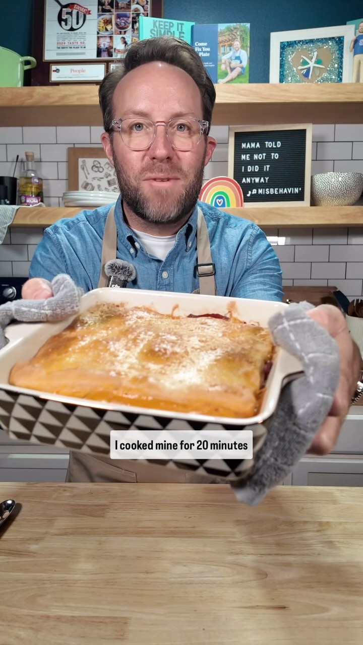 A man with glasses holds a casserole dish towards the camera, with a white casserole in front of him, wearing an apron. Text at the bottom says 'I cooked mine for 20 minutes'. The background includes a tiled wall, shelves with books, framed artwork, decor, and letterboard.