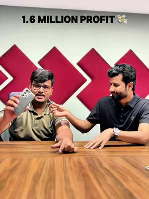 Two men are seated at a wooden table. The man on the left is holding up a phone and wearing glasses; the man on the right is gesturing toward the phone. In the background, there are red art pieces on a wall.