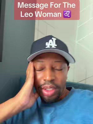 A person wearing a cap and looking at the camera with a slight smile is a top-down shot. Overlay text is present at the top, reading 'Message for the Leo Woman'. The background is a mix of grey and blue. The person is wearing a dark blue cap with the letters A and L on it, and a blue shirt.