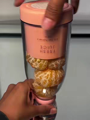 A person is holding a clear cylindrical container, possibly a juicer or blender, filled with peeled orange segments. The container is clear, allowing the orange color to be visible. The container has a pink cap and base, and a pink band with text that reads 'FRESH JUICE'. The person's hands are visible, with dark skin and the person's fingers can be seen holding and operating the juicer. A white surface acts as the background. The overall shot highlights the juicer and its contents..