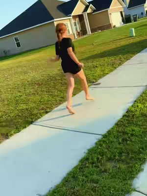 A woman with red hair is barefoot, walking down a sidewalk. The background includes green grass, houses, and a bright sky.