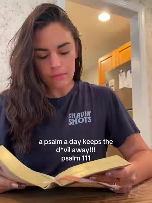 A woman is seated and holding a Bible open. She looks down toward the book, and the lighting suggests she's indoors. The background is slightly blurred, including parts of a kitchen area.