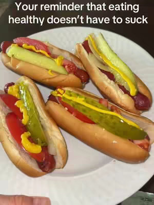 A plate of four hot dogs are presented. The hot dogs are arranged side-by-side, with their filling ingredients, including mustard, pickle slices, and tomato, adding vibrant color. The image is bright and well-lit.