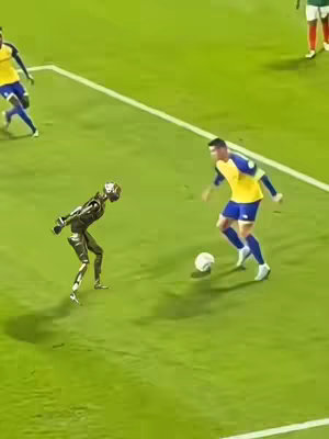 The image shows a soccer field with a robot and a soccer player. The robot is black and silver and is posed as if it is about to play the ball. The soccer player is wearing a yellow and blue uniform, is standing with the ball and performing a soccer move. The background shows the soccer field with other players on the field.