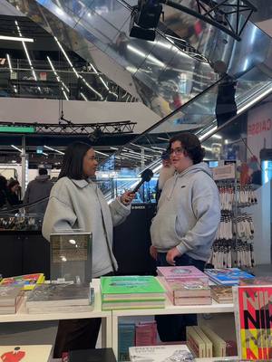 Two people are shown in a retail setting, with a woman on the left holding a microphone and appearing to interview a person on the right. They are surrounded by shelves of books and other items. The background features a shiny ceiling with lights and a staircase.