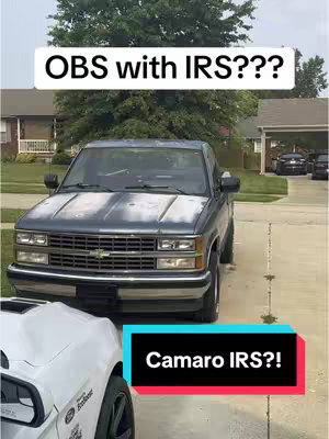 The image shows a dark gray pickup truck parked on a driveway, with two lines of text overlaid. The truck has a weathered appearance, and a white car's side is visible in the frame's lower left corner. In the background, a house and trees are partially visible. The overall composition is straightforward, emphasizing the truck against a residential backdrop.