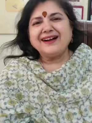 A woman with dark hair and a small bindi on her forehead smiles at the camera. She wears a patterned top and is positioned against a blurred background.