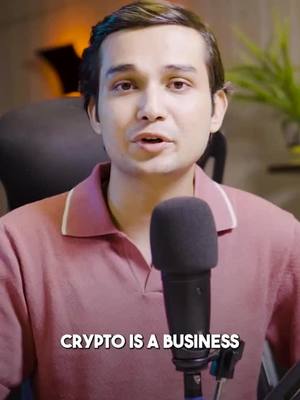 A man with short brown hair is centered in frame, speaking into a microphone. He wears a pink polo shirt with a white collar. The bottom of the image bears the text 'CRYPTO IS A BUSINESS' in a white, sans serif font. Behind him is a mostly blurred-background with some office-like, dimly lit setting.