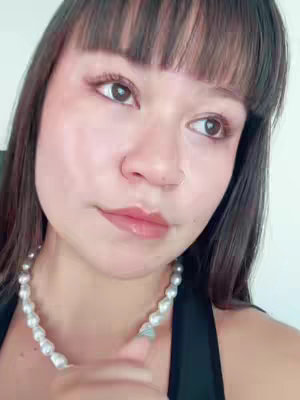 A close-up shot of a woman with dark hair and bangs, looking away from the camera with a thoughtful expression. She wears a pearl necklace and a black top against a neutral background.
