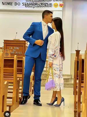 A couple formally dressed in a church setting. The man, in a blue suit, is leaning in to kiss the woman's forehead. The woman is wearing a long dress with floral patterns and a small purse with matching color of the heels.