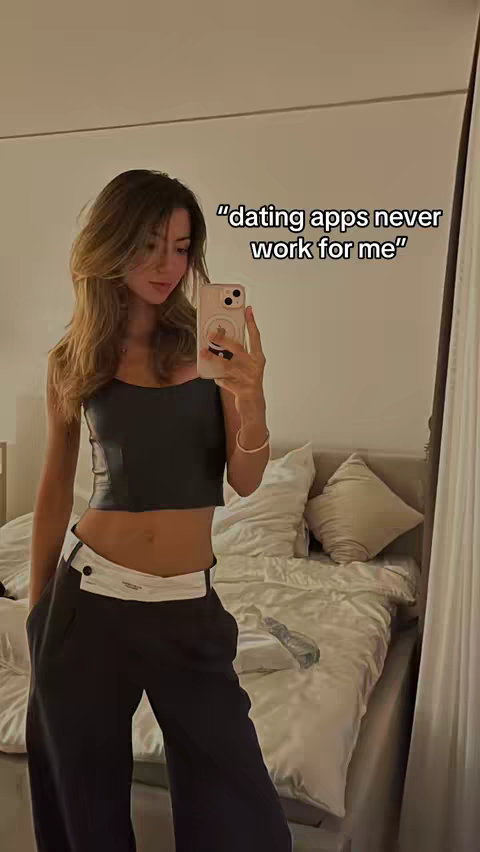 A young woman in a stylish outfit is taking a mirror selfie. She is wearing a black crop top and black pants with a white waistband. The background shows a bedroom with a bed covered in white sheets, a mirror, and a window.
