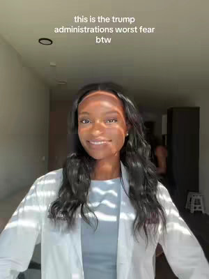 A young woman of color stands in a well-lit hallway, smiling at the camera. She is wearing a white lab coat over a light blue top. The background includes a dark doorway, a ceiling light, and a glimpse of another person in the distance. The lighting suggests sunlight, casting shadows and highlights on her face and the surrounding area. The main text overlay is in white.