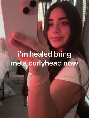 A young woman is holding a phone to take a selfie; she is wearing a white top and a bracelet, with long dark hair, and is looking towards the camera with a slight smile. Text overlayed on the image reads, “I’m healed bring me a curlyhead now.” The image is lit with a reddish hue. A white bed is visible in the background.