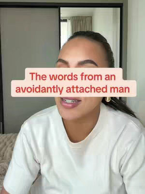 A selfie-style shot featuring a woman with dark hair, wearing a white t-shirt. The background shows an off-white wall, a mirror, and a glimpse of window or door. A rectangular text overlay partially obscures her face. The text is pink and reads, 'The words from an avoidantly attached man'. The woman has her mouth slightly open, showcasing her braces.