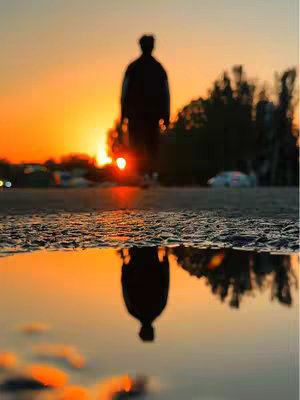 A backlit silhouette of a person walking towards the viewer, reflecting in a puddle on the ground. The sky is vibrant with an orange and yellow sunset. The composition is symmetrical with the reflection mirroring the scene above.