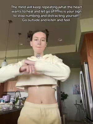 The image is a selfie-style shot of a person indoors, likely in their home. The individual, with light skin and dark hair styled in space buns, is positioned centrally in the frame and wearing a cream-colored hoodie and matching sweatpants. The background features a kitchen with cabinets, appliances, and some decor that add to the homey feel. Text overlay on the image provides self-help advice.