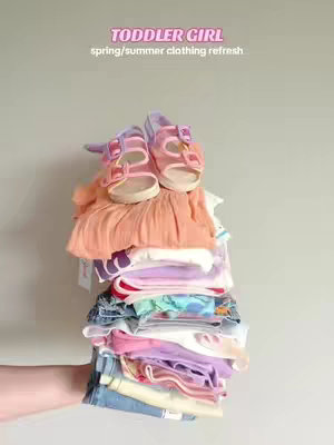 A hand holds a tall stack of colorful toddler clothing, with a pair of pink and purple sandals resting on top. The clothes include a variety of tops, pants, and dresses in pastel and bright colors. A light gray-green background frames the image.