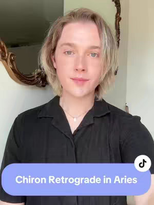 The cover image is a selfie of a person with short, light blonde hair, wearing a black collared shirt. Behind the person is a wall and a decorative gold mirror. A text overlay in a light blue rectangle reads "Chiron Retrograde in Aries". The light is natural looking.