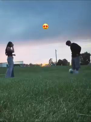 A woman and a man are standing on a grassy field. The woman, in the left, is facing the camera with her hand up to her face, while the man, on the right, is bending down, seemingly about to kick a soccer ball with an emoji above their heads. The sky is a mix of dusk colors.