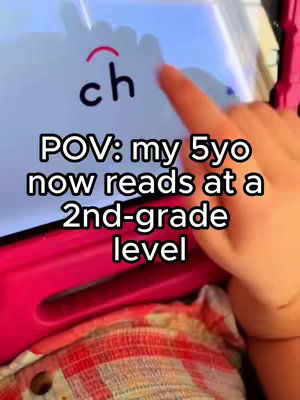A person's hand points to a tablet screen displaying the letters 'ch' with a diacritic mark. overlaid text says 'POV: my 5yo now reads at a 2nd-grade level'. The background contains parts of a pink case and a patterned blanket.