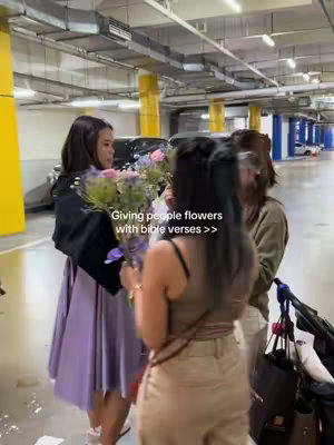 Three young women in a parking garage, exchanging flowers. One woman on the left wears a purple dress and holds a bouquet, the central woman is in a beige top holding flowers, with the third woman partially visible on the right. The background is a typical parking garage with yellow support pillars and overhead lighting.
