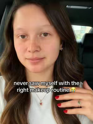 A woman is in the front seat of a car, facing the camera. She has long, dark hair and is wearing a white shirt and a delicate necklace with a pendant. In the foreground, her hand with red fingernails is visible. The background includes parts of the car’s interior and a blurred view of the outside.