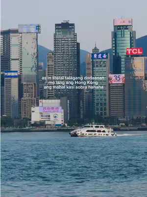 The cover image shows a scenic view of Hong Kong's skyline and harbor. The foreground is dominated by the deep blue water, with a white ferry boat moving from left to right. The background features a densely concentrated cityscape with numerous skyscrapers of different heights and designs, some with illuminated signs. The sky is a gradient of colors from pink to blue, suggesting a dawn or dusk setting. Text overlays in both English and Chinese cover the image.