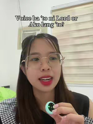 A young woman with fair skin, wearing glasses and a black top, holds a small mint-green device, likely a microphone, in her hand. The background includes a light-colored wall, a window with blinds, and possibly a glimpse of a green object. The text overlay at the top asks: "Voice ba 'to ni Lord or Ako lang 'to?"