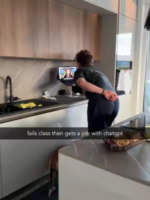 A person is standing in a kitchen, facing a laptop on a stand on a counter. The person's back is to the camera as they watch the screen, and the kitchen has light colored cabinets and countertops.