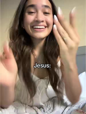 A young woman with long brown hair, wearing a white top, smiles widely at the camera with her hands up as if offering something. The text overlay says "Jesus:"