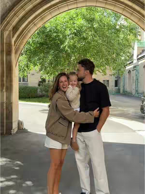 A family of three is captured in a warm embrace under an archway, suggesting a moment of togetherness. The scene is filled with natural light and greenery, creating a soft, joyful atmosphere. The color palette is muted which creates a sense of peace; the composition suggests a family who is affectionate and happy.
