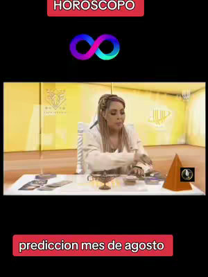 The image shows a woman in a seated position, presumably in a studio, with a table in front of her. Various items are on the table, including tarot cards, a small golden item, and a pyramid-shaped object. Text overlays at the top and bottom read, "HOROSCOPO" and "prediccion mes de agosto," respectively. An infinity symbol is in the middle. The background is a soft gradient of yellow/gold, with the overall tone being one of mysticism and the esoteric. The woman is fair-skinned and has her hair pulled back.