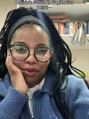 A woman with dark hair and glasses is looking at the camera and touching her face with her hand. Text overlayed at the top says 'Cassie's Saturn in Sagittarius 7H!?.' The background is blurred.