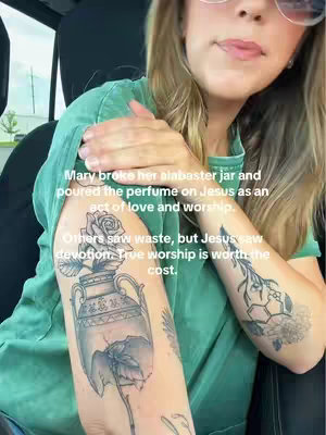A woman wearing a green top is holding her arm up, showcasing two tattoos. On her bicep, there's a large tattoo of a vase with a rose, broken to show the inside. The other arm has another tattoo on the forearm, featuring a geometric design. Text overlays the image, discussing Mary and worship, linking to the tattoos.