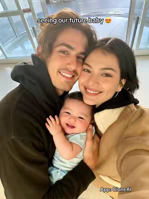 A selfie-style image features a smiling couple, a man with dark hair and a mustache on the left and a woman on the right, both holding a baby in the center of the frame. The baby is also smiling. The woman's arm is around the baby.