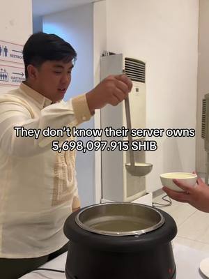A man in a light-colored shirt is shown serving soup. The scene is set in a dining area with a sign directing to restrooms and elevators. Text overlay states, "They don't know their server owns 5,698,097.915 SHIB."