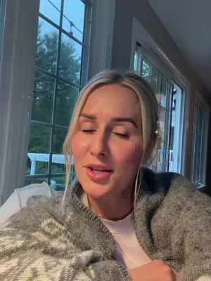 A woman with blonde hair and a sweater is sitting in what appears to be a living room, with a window behind her. The woman has her eyes closed and seems to be speaking. The overall image has a sense of comfort and relaxation.